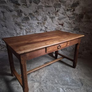 Peut inclure: Une table rectangulaire en bois au fini brun naturel, dotée d'un tiroir unique équipé d'une poignée en anneau en métal foncé. La table est soutenue par quatre pieds droits reliés par une barre transversale horizontale près de la base.