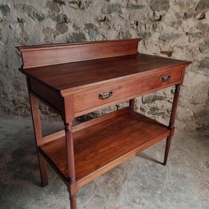 Op de afbeelding: Een bruine houten wastafel met een rechthoekig blad en een opstaande achterkant, een brede lade met twee sierlijke metalen handgrepen en een open onderste plank, ondersteund door vier poten met verticale groeven.