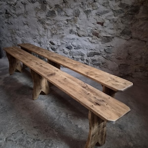 Op de afbeelding: Twee lange, smalle houten banken met een natuurlijke lichtbruine afwerking met zichtbare houtnerven en knoesten. Elke bank wordt ondersteund door twee stevige, wijd uit elkaar geplaatste poten met een uitsparing aan de onderkant.