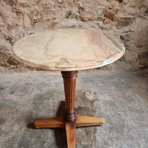 Op de afbeelding: Een ronde tafel met een gepolijst marmeren blad met wervelende roze, witte en grijze aders. De tafel wordt ondersteund door een centrale houten kolom met verticale groeven, rustend op een vierpotige, kruisvormige houten basis.