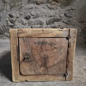 Op de afbeelding: Een kleine, rechthoekige houten kast met een verweerde, natuurlijk bruine afwerking. De voorzijde is voorzien van een enkele draaideur met een donker metalen sleutelgatplaatje en een zichtbaar metalen scharnier aan de rechterkant van het hout.