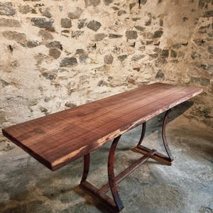 Op de afbeelding: Een rechthoekige houten tafel met een natuurlijke rand en een roodbruine afwerking. De tafel wordt ondersteund door gebogen, roest-rode metalen poten en een rechthoekige basis. De tafel is ongeveer 183 cm lang.