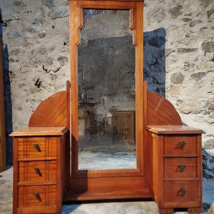 Op de afbeelding: Een vintage houten kaptafel met een grote rechthoekige spiegel en twee zijkasten, elk met drie lades. De kaptafel is gemaakt van warm getint hout en heeft decoratieve details. De spiegel is omlijst door een gedetailleerde houten rand. De kaptafel staat tegen een stenen muur.