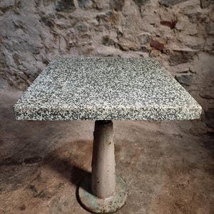Op de afbeelding: Kleine, vierkante tafel met een gespikkelde grijze granieten bovenkant. De tafel wordt ondersteund door een lichtgrijze, cilindrische voet met een ronde voet. Het ontwerp van de tafel is eenvoudig en functioneel.