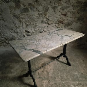 Peut inclure: Une table rectangulaire dotée d'un plateau en marbre blanc aux veines grises marquées et aux coins arrondis. La table repose sur une base en métal noir composée de deux pieds ornés reliés par une barre de support horizontale.