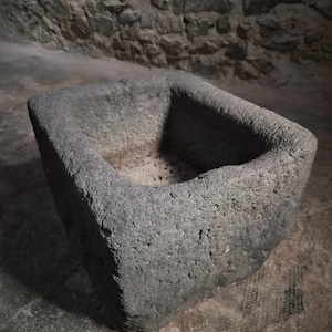 Könnte beinhalten: Ein schweres, rechteckiges Steinbecken mit einer rauen, porösen grauen Textur. Das Gefäß verfügt über dicke, massive Wände und eine tiefe, ausgehöhlte Mitte, die ein natürliches, verwittertes Aussehen mit sichtbaren Vertiefungen aufweist.