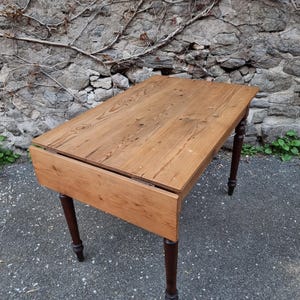 Peut inclure: Une table rectangulaire en bois à abattant dotée d'un plateau au grain brun clair et de quatre pieds tournés brun foncé. La table possède un panneau latéral articulé qui peut être rabattu, révélant une finition naturelle avec des nœuds.