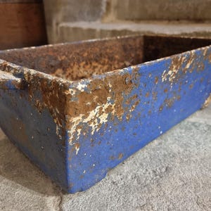 Op de afbeelding: Rechthoekige blauwe metalen container met handvatten, met tekenen van roest en slijtage. De container heeft een verweerde uitstraling met afbladderende verf en een verweerde textuur, wat duidt op leeftijd en gebruik. De container wordt waarschijnlijk gebruikt voor opslag.