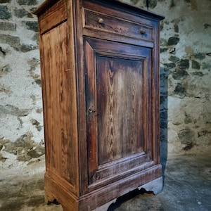 Peut inclure: Un meuble en bois ancien, avec une porte et un tiroir. Le meuble est en bois teinté foncé avec un grain visible et comporte un petit bouton rond sur le tiroir. Le meuble est placé contre un mur en pierre.