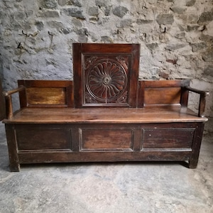 Könnte beinhalten: Eine dunkelbraune Holzbank mit einer hohen Rückenlehne, die in der Mitte ein quadratisches Paneel mit einem geschnitzten kreisförmigen Blumenmotiv aufweist. Die Bank hat flache Armlehnen und eine rechteckige Sitzfläche mit drei eingelassenen Paneelen.