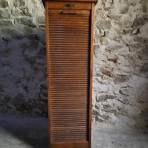 Könnte beinhalten: Ein hoher, rechteckiger Holzschrank mit einer vertikalen Rollladentür aus horizontalen Lamellen. Der Schrank hat eine dunkelbraune Holzmaserung, eine flache Oberseite und einen kleinen Metallgriff, der im oberen Bereich der Tür eingelassen ist.