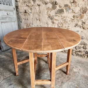 Op de afbeelding: Een ronde, lichtbruine houten tafel met een klepdesign. De tafel heeft vier poten en een natuurlijke houtnerf afwerking. De tafel heeft een diameter van ongeveer 100 cm wanneer volledig uitgeklapt.