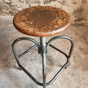 Op de afbeelding: Een vintage houten kruk met een ronde zitting en een metalen frame. De zitting heeft een versleten uitstraling, met zichtbaar hout en afbladderende verf. De metalen basis is donkergrijs en heeft een uniek, geometrisch ontwerp. De zitting is ongeveer 30 cm in diameter.