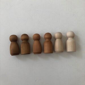 May include: Seven wooden peg dolls in a row, painted in various shades of brown and white. The dolls are all the same size and shape, with a round head and a simple body.