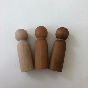 May include: Three wooden peg dolls, one is natural wood, one is brown, and one is a darker brown.