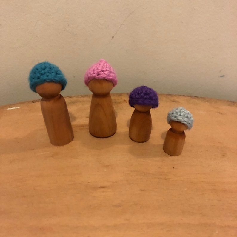 May include: Four wooden peg dolls wearing crocheted hats in different colors: blue, pink, purple, and light blue.
