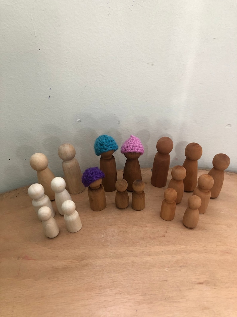 May include: A collection of wooden peg dolls, some wearing crocheted hats in purple, pink, and blue. The dolls are arranged on a wooden surface.
