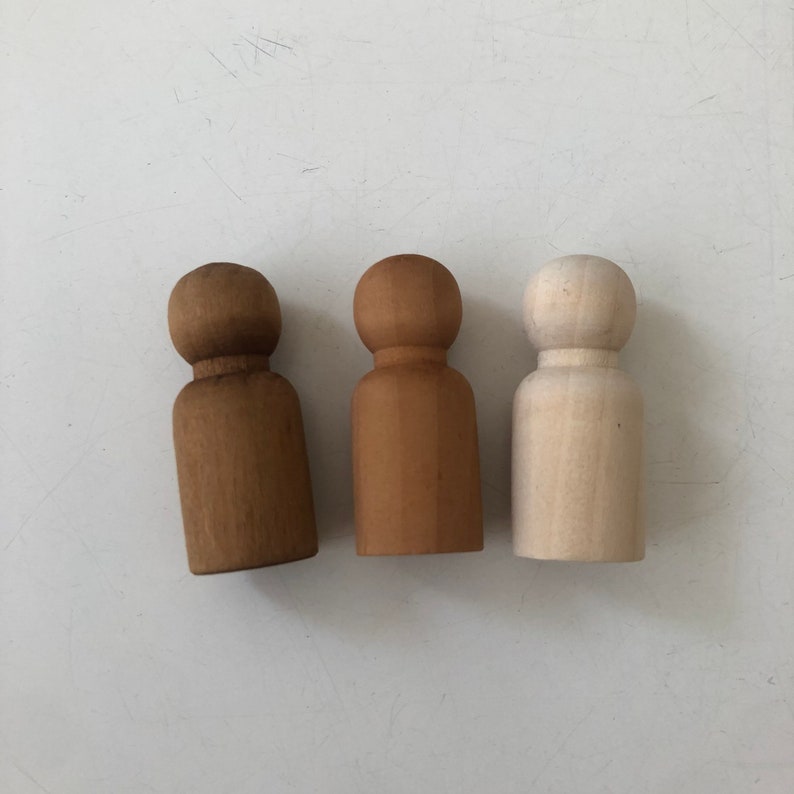 May include: Three wooden peg dolls, one is natural wood, one is stained a light brown, and one is stained a darker brown.