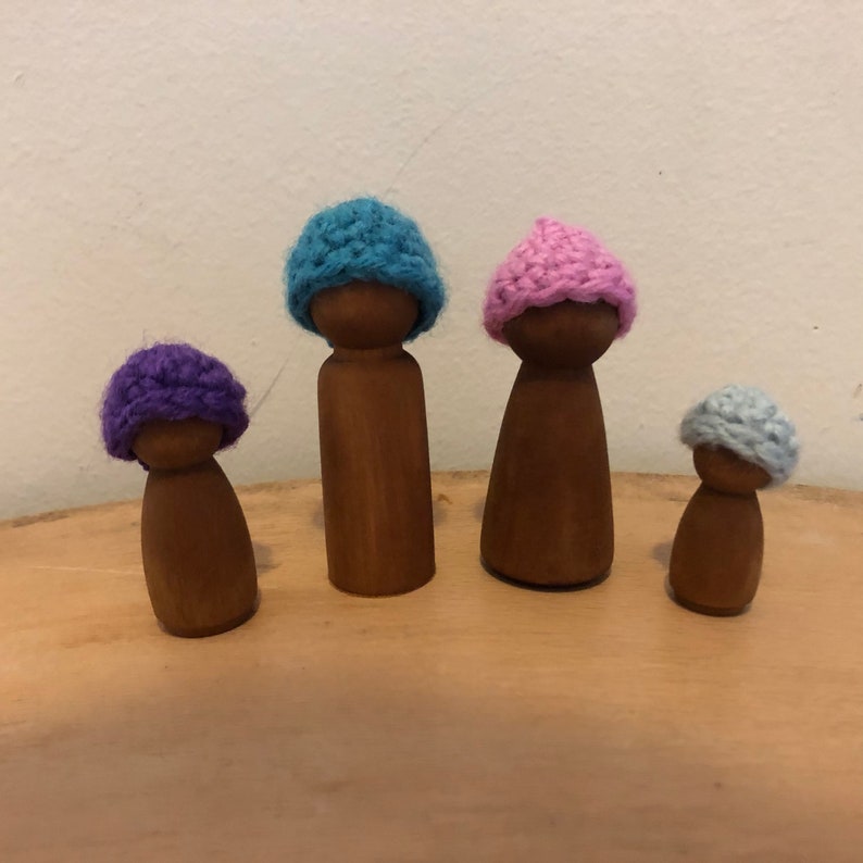 May include: Four wooden peg dolls wearing crocheted hats. The hats are in shades of blue, pink, purple, and light blue.