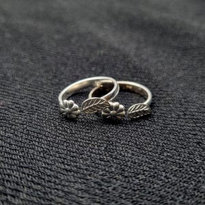 Könnte beinhalten: Zwei silberne Zehenringe mit floralen und Blattmotiven. Jeder Ring hat eine kleine Blume und einen Blattschmuck. Die Ringe werden auf einem dunklen, strukturierten Hintergrund präsentiert, der die filigranen Details des Schmucks hervorhebt.