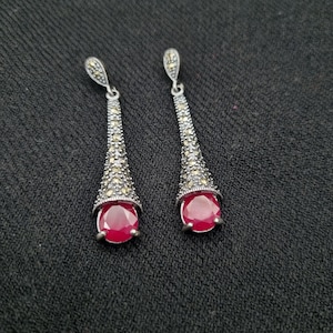 May include: A pair of silver-toned drop earrings. Each earring features a teardrop-shaped design with a red gemstone at the base. The earrings are embellished with small, sparkling accents and have a hook closure.