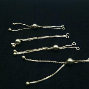 Op de afbeelding: Close-up van verschillende zilverkleurige verstelbare armbandkettingen. Elke ketting heeft een kleine bal en een verschuifbare kraal voor aanpassing. De kettingen worden weergegeven tegen een donkere achtergrond, wat hun metaalglans benadrukt.