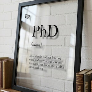 Puede incluir: Impresión enmarcada en negro con la palabra "PhD" en negrita. Debajo, la definición dice: "un académico que ha aprendido más y más sobre menos y menos hasta que lo saben todo sobre nada."
