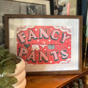 May include: Framed art print with the words "FANCY PANTS" in pink and teal lettering. The print features illustrations of underwear hanging on a clothesline against a red background. The frame is brown.