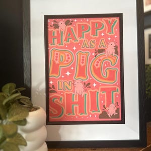 May include: Framed art print with the phrase "HAPPY AS A PIG IN SHIT" in colorful lettering. The print features cartoon pigs and a coral background, framed in black with a white mat.
