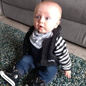 May include: A baby wearing a black and white striped sweater, blue jeans, and black high-top trainers with white soles. The baby is sitting on a green and white patterned rug.