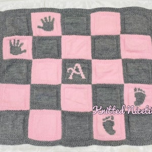 May include: A pink and grey knitted baby blanket with hand and foot prints and the letter "A" in the centre. The blanket has the text "Knitted Niceties" in the bottom right corner.