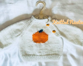 May include: White knitted baby sweater with an orange pumpkin design and yellow buttons. The sweater has the text "Knitted Niceties" on the right side.
