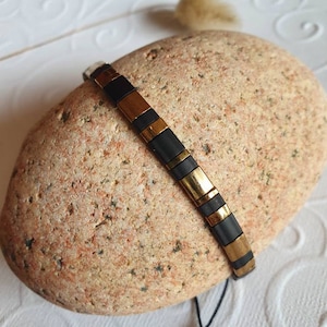 Peut inclure: Un bracelet noir et or composé de petites tuiles carrées. Le bracelet est posé sur une pierre brun clair.