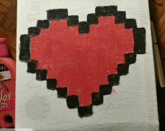 May include: A white canvas with a large, pixelated red heart outlined in black. The heart design is centered on the canvas. The bottom left corner has the text "NERDY RENDITION" with icons. The canvas is on a wooden surface.