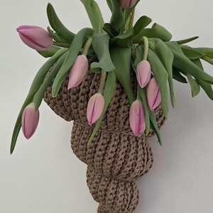 Könnte beinhalten: Ein brauner, gehäkelter Wandtopf in Muschelform, gefüllt mit rosa Tulpen und grünen Blättern. Die Tulpen befinden sich in verschiedenen Blühstadien. Ein hölzernes Herz schmückt die Wand.