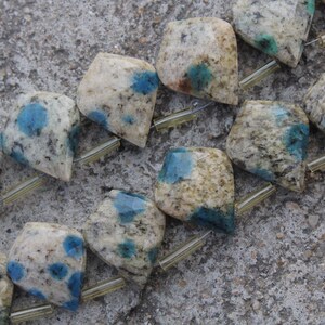 Può includere: Primo piano di una collana di perline di pietra K2. Le perline sono un mix di bianco, grigio e beige, con macchie blu e verdi. Le perline sono infilate su un cordino sottile e trasparente.