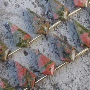 Puede incluir: Primer plano de cuentas de piedra preciosa Unakita. Cada cuenta triangular tiene una varilla de metal dorada. Las cuentas de Unakita muestran una mezcla de colores verdes, rojos y marrones. Para joyería.