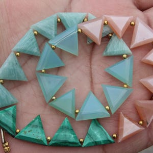 Peut inclure: Un collier composé de perles triangulaires en pierres précieuses vertes, bleues et pêche. Les perles sont reliées par de petits accents dorés. Le collier est présenté sur une main.