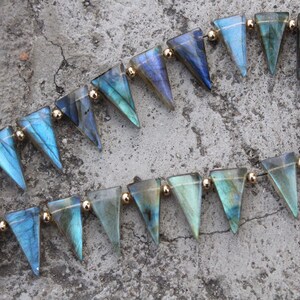 Op de afbeelding: Twee strengen edelsteen kralen. Elke kraal heeft een driehoekige vorm met blauwe en groene iriserende kleuren. De kralen zijn geregen met goudkleurige kralen tussen elke edelsteen.