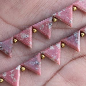 May include: Close-up of two strands of pink triangular gemstone beads with gold-colored spacers. The gemstones have a mottled appearance with darker pink and white inclusions. The beads are arranged in a row, creating a geometric pattern.
