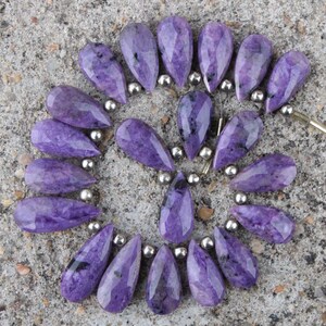 Puede incluir: Una colección de cuentas de charoíta moradas, facetadas y con forma de pera, dispuestas en un patrón circular. Cada piedra preciosa presenta inclusiones oscuras naturales y está separada por pequeñas cuentas espaciadoras metálicas plateadas.