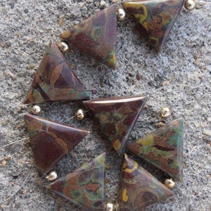 Op de afbeelding: Een ketting met driehoekige kralen in bruine, groene en gele tinten, verbonden door goudkleurige kralen. Het geometrische ontwerp van de kralen creëert een unieke en aardse esthetiek, geschikt voor diverse outfits.