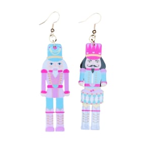 May include: A pair of dangle earrings featuring a pink and blue nutcracker design. The earrings are made of acrylic and have gold hooks.