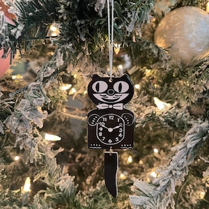 May include: Black and white cat clock ornament with a swinging tail. The clock face has black hands and white numbers.