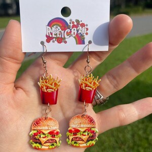 May include: Dangling earrings featuring a red container of fries and a burger. The burger has a sesame seed bun, lettuce, tomato, cheese, and a beef patty. The earrings are attached to a white tag with the text "RELIC828" and a rainbow design.