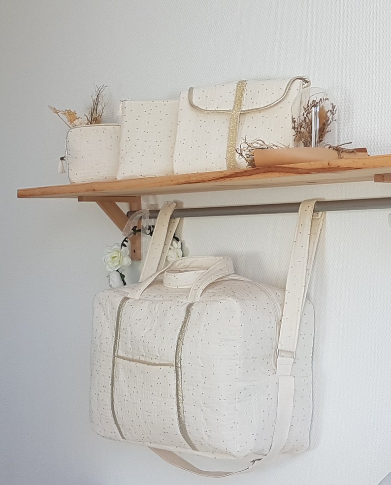 May include: A set of four off-white canvas bags with gold accents. The bags are lined up on a wooden shelf. The largest bag has two shoulder straps and a top handle. The smaller bags have a zipper closure.