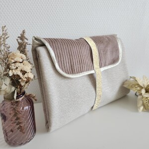 May include: A beige clutch bag with a textured, glittery fabric. The flap is a ribbed, mauve material, outlined in white. A gold ribbon secures the flap. A vase of dried flowers and a gold decorative flower are in the background.