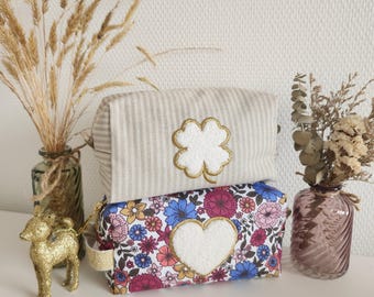 May include: Two cosmetic bags. The top bag is beige with vertical stripes and a white four-leaf clover with gold trim. The bottom bag has a floral pattern with a white heart. Decorative vases and a gold deer figurine are also present.