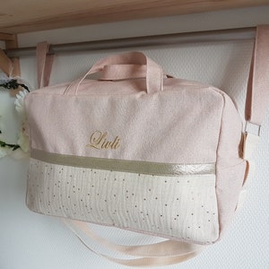 May include: A pink and white canvas bag with a gold trim and gold polka dots. The bag has a long strap and a handle. The name "Liuli" is embroidered on the front of the bag.