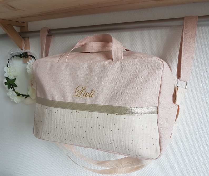 May include: A pink and white canvas bag with a gold trim and gold polka dots. The bag has a long strap and a handle. The name "Liuli" is embroidered on the front of the bag.